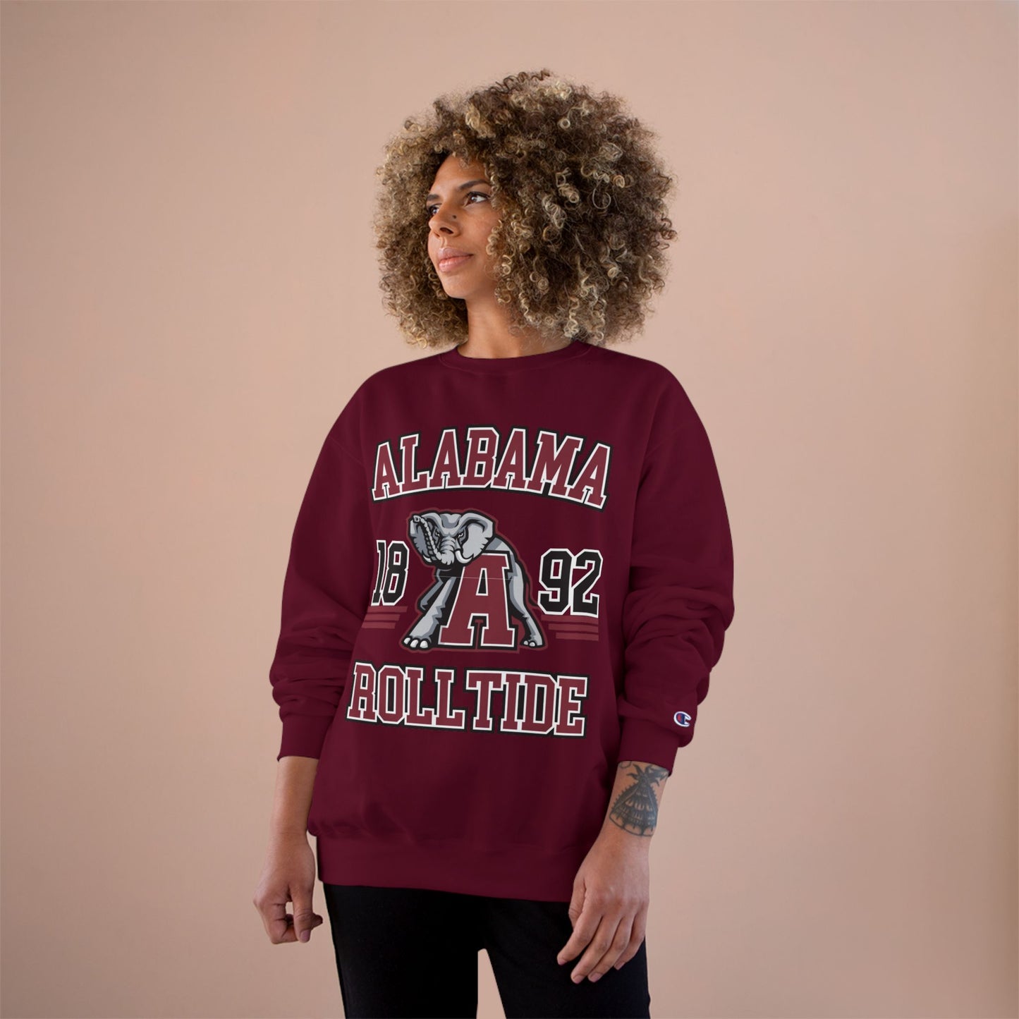 Person wearing a maroon 'Alabama Roll Tide' sweatshirt with a logo on a beige background